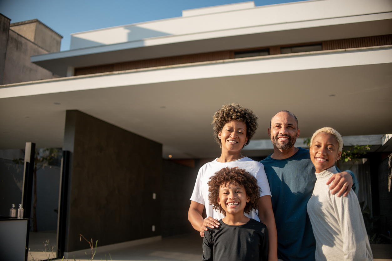 This image shows a black family with a dad, mom, daughter and son smiling and standing in front of their supportive housing building.