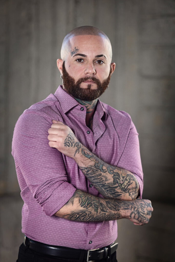 Man with tattoos wearing purple collared shirt. 
