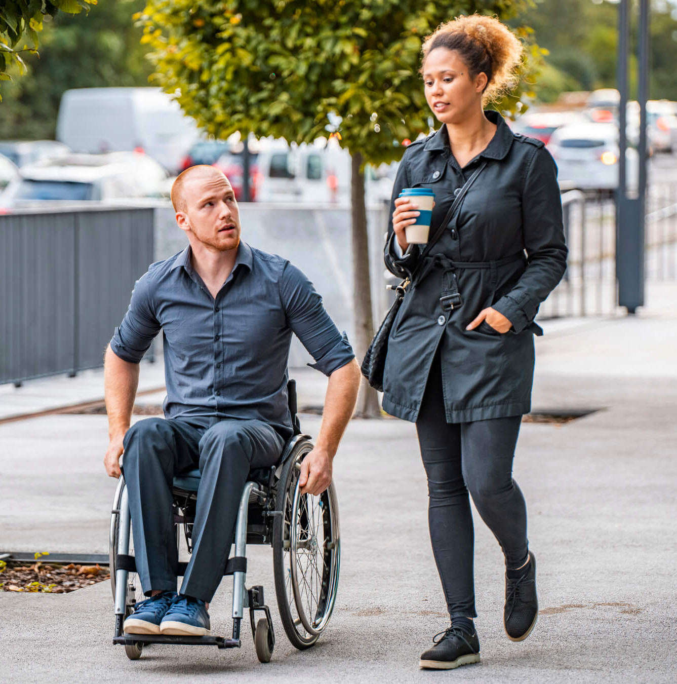 A woman and man are walking down an urban street, having a conversation. The woman is holding a coffee cup and the man is in a wheelchair. 