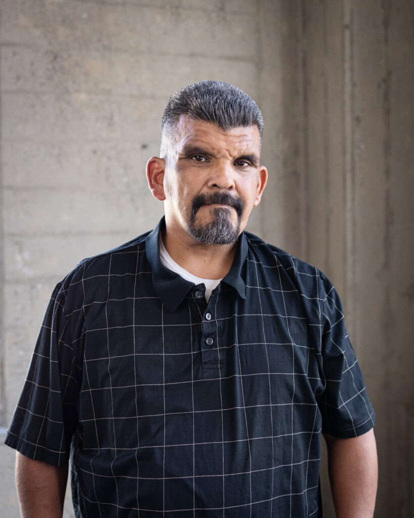 smiling man standing in front of a wall wearing a button down shirt 