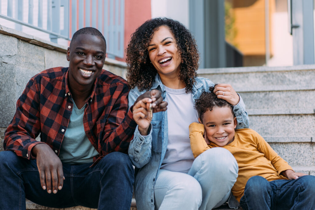 Portrait of happy family with one child moving to new house . A young family move into their new home. Family of three sitting in front the house and proudly displays the keys to their new home.